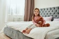 A cute little browneyed girl sitting barefoot on a huge bed holding her teddybear with her feet crossed wearing pyjamas