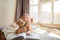 Cute little caucasian girl in casual clothes reading a book with stuffed teddy bear toy and smiling while lying on a Royalty Free Stock Photo