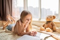 Cute little caucasian girl in casual clothes reading a book with stuffed teddy bear toy and smiling while lying on a floor near Royalty Free Stock Photo