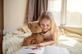Cute little caucasian girl in casual clothes reading a book with stuffed teddy bear toy and smiling while lying on a floor near Royalty Free Stock Photo