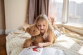 Cute little caucasian girl in casual clothes reading a book with stuffed teddy bear toy and smiling while lying on a floor near Royalty Free Stock Photo