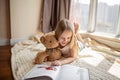 Cute little caucasian girl in casual clothes reading a book with stuffed teddy bear toy and smiling while lying on a Royalty Free Stock Photo