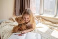 Cute little caucasian girl in casual clothes reading a book with stuffed teddy bear toy and smiling while lying on a Royalty Free Stock Photo