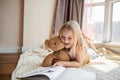 Cute little caucasian girl in casual clothes reading a book with stuffed teddy bear toy and smiling while lying on a Royalty Free Stock Photo