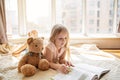 Cute little caucasian girl reading a book with stuffed teddy bear toy. Stay at home during coronavirus covid-19 pandemic Royalty Free Stock Photo