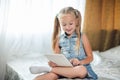 Cute little child girl blond in denim sundress lies in bed uses digital tablet. child playing on tablet pc having fun and smiles