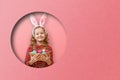 Cute little child girl with bunny ears holding basket of Easter eggs. Child in a round hole circle in colored pink background Royalty Free Stock Photo