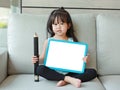 Cute little child girl holding empty white blackboard with big pencil sitting on fabric sofa in library room. Education concept Royalty Free Stock Photo