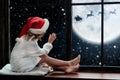 Cute little child sitting by window and looking at Santa Claus flying in his sleigh against moon sky Royalty Free Stock Photo