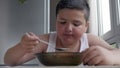 Cute little fat boy dining in kitchen, eating a tablespoon of soup, concept childhood obesity and gluttony