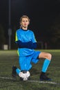 cute little footballer girl kneeling in a stadium with a soccer ball, vertical full shot Royalty Free Stock Photo