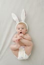 Cute little funny caucasian baby girl wearing white rabbit ears and diaper lying on back mouthing foot looking at camera Royalty Free Stock Photo