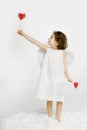 Cute little girl in angel look, in white dress and wings, holding a red heart in hands, looking in profile. Royalty Free Stock Photo