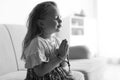 Cute little girl with beads praying on sofa in living room, black and white effec Royalty Free Stock Photo