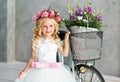 A cute little girl in a beautiful lush white dress in a wreath of flowers on her head. standing in the studio next to the bike Royalty Free Stock Photo