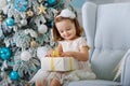 Cute little girl in bklom dress sitting in a chair and opens box with present for background Christmas tree blue Royalty Free Stock Photo