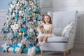Cute little girl in bklom dress sitting in a chair and opens box with present for background Christmas tree blue Royalty Free Stock Photo