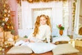 Cute little girl with blond long curly hair in a light knitted sweater by the festive fireplace with garlands Royalty Free Stock Photo