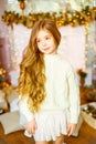 Cute little girl with blond long curly hair in a light knitted sweater by the festive fireplace with garlands Royalty Free Stock Photo