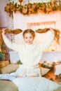 Cute little girl with blond long curly hair in a light knitted sweater by the festive fireplace with garlands Royalty Free Stock Photo