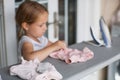 Cute little girl with blonde hair is leaning on ironing clothes on board at home. Daughter helping to mother ironing clothes for Royalty Free Stock Photo