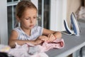 Cute little girl with blonde hair is leaning on ironing clothes on board at home. Daughter helping to mother ironing clothes for Royalty Free Stock Photo