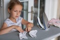 Cute little girl with blonde hair is leaning on ironing clothes on board at home. Daughter helping to mother ironing clothes for Royalty Free Stock Photo