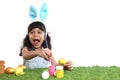 Cute little girl with blue bunny ears painting eggs with brush on green grass meadow, joyful kid celebrating Easter holiday on Royalty Free Stock Photo