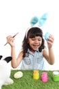 Cute little girl with blue bunny ears painting eggs with brush on green grass meadow, joyful kid celebrating Easter holiday on Royalty Free Stock Photo