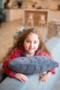 Cute little girl with blue eyes and long hair in red pajamas lies on gray sofa in living room, hugs a pillow and smiles Royalty Free Stock Photo