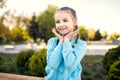 A cute little girl in a blue jacket with pigtails poses while sitting on a bench with her hands pressed to her face Royalty Free Stock Photo