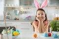 Cute little girl with bunny ears headband and painted Easter eggs sitting at table in kitchen Royalty Free Stock Photo