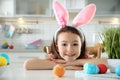 Cute little girl with bunny ears headband and painted Easter eggs sitting at table in kitchen Royalty Free Stock Photo