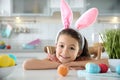 Cute little girl with bunny ears headband and painted Easter eggs sitting at table in kitchen Royalty Free Stock Photo