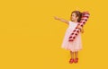 A cute little girl child in a fancy dress and a Santa hat holds an inflatable shape of a candy cane on a yellow background. 2021 Royalty Free Stock Photo