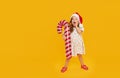 A cute little girl child in a fancy dress and a Santa hat holds an inflatable shape of a candy cane on a yellow background. 2021 n Royalty Free Stock Photo
