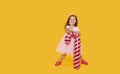 A cute little girl child in a fancy dress and a Santa hat holds an inflatable shape of a candy cane on a yellow background. 2021 Royalty Free Stock Photo