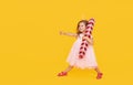 A cute little girl child in a fancy dress and a Santa hat holds an inflatable shape of a candy cane on a yellow background. 2021 Royalty Free Stock Photo