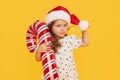 A cute little girl child in a fancy dress and a Santa hat holds an inflatable shape of a candy cane on a yellow background. 2021 Royalty Free Stock Photo