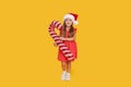 A cute little girl child in a fancy dress and a Santa hat holds an inflatable shape of a candy cane on a yellow background. 2021 Royalty Free Stock Photo