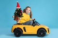 Cute little girl with Christmas tree driving children`s electric toy car on light blue background Royalty Free Stock Photo