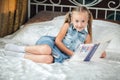 Cute little girl in denim sundress reading book looking at camera and smiling on bed at home Royalty Free Stock Photo