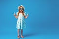 Cute little girl in a dress, hat and sunglasses poses on a blue background