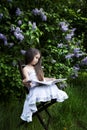 Cute little girl, dressed in a white dress, sitting in the garden in the park and reading a book. green and blooming lilacs Royalty Free Stock Photo