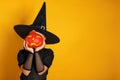 Cute little girl dressed in a witch costume holds a pumpkin instead of a head on a yellow background. Halloween celebration Royalty Free Stock Photo