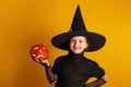 Cute little girl dressed in a witch costume holds a pumpkin jack lantern on a yellow background. Halloween celebration Royalty Free Stock Photo