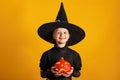 Cute little girl dressed in a witch costume holds a pumpkin jack lantern on a yellow background. Halloween celebration Royalty Free Stock Photo