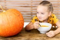 Cute little girl eating pumpkin soup and looking at a large Halloween pumpkin, with vicious face expression. Halloween.