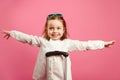 Cute little girl extended arms, depicts a plane, smiling happily, stands near suitcase on pink.