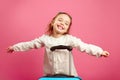 Cute little girl extended arms, depicts a plane, smiling happily, stands near suitcase on pink.